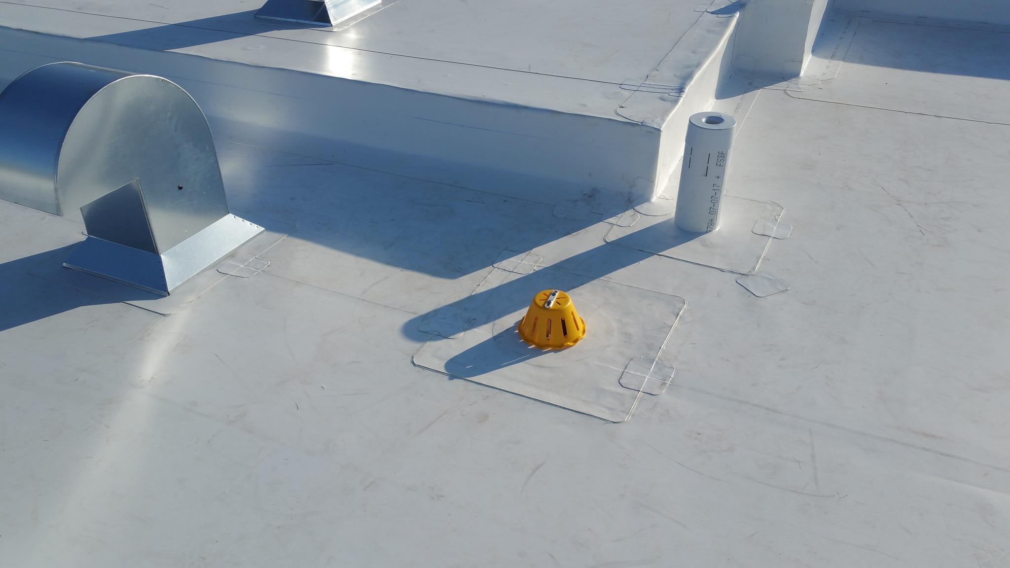 Détail d'un toit plat recouvert d'une membrane blanche, montrant des conduits de ventilation et un drain de toit jaune, assurant une bonne gestion de l'eau et la ventilation du bâtiment.