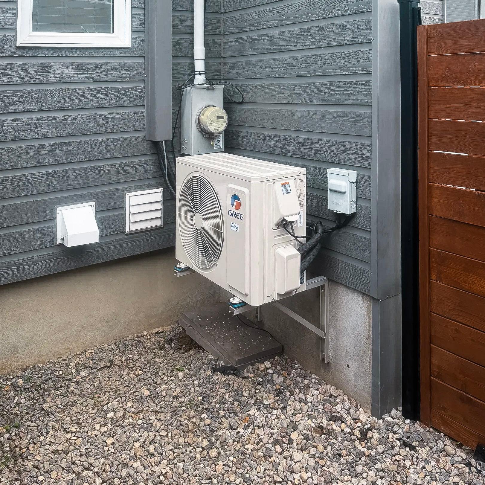 Unité extérieure d'un climatiseur de marque Gree installée sur un mur extérieur, avec un revêtement gris et un sol en gravier, à côté d'une fenêtre et d'une clôture en bois.<br>