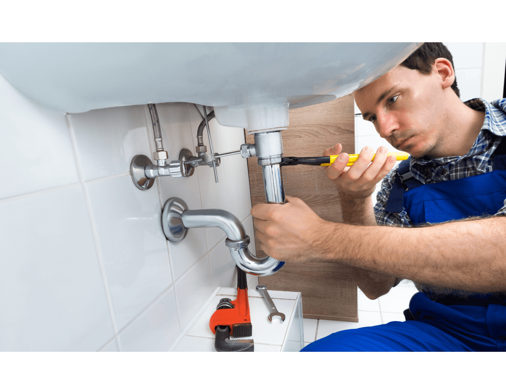 Plombier en salopette bleue réparant un siphon de lavabo avec une clé, dans une salle de bain carrelée.
