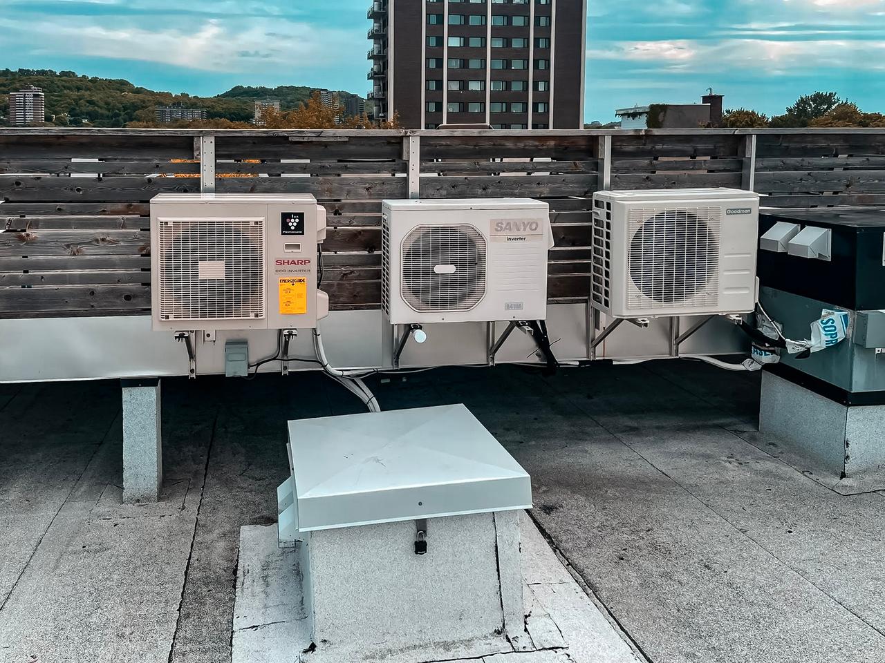 Unités de climatisation extérieures installées sur un toit plat avec vue sur des immeubles résidentiels et collines verdoyantes à l'arrière-plan.