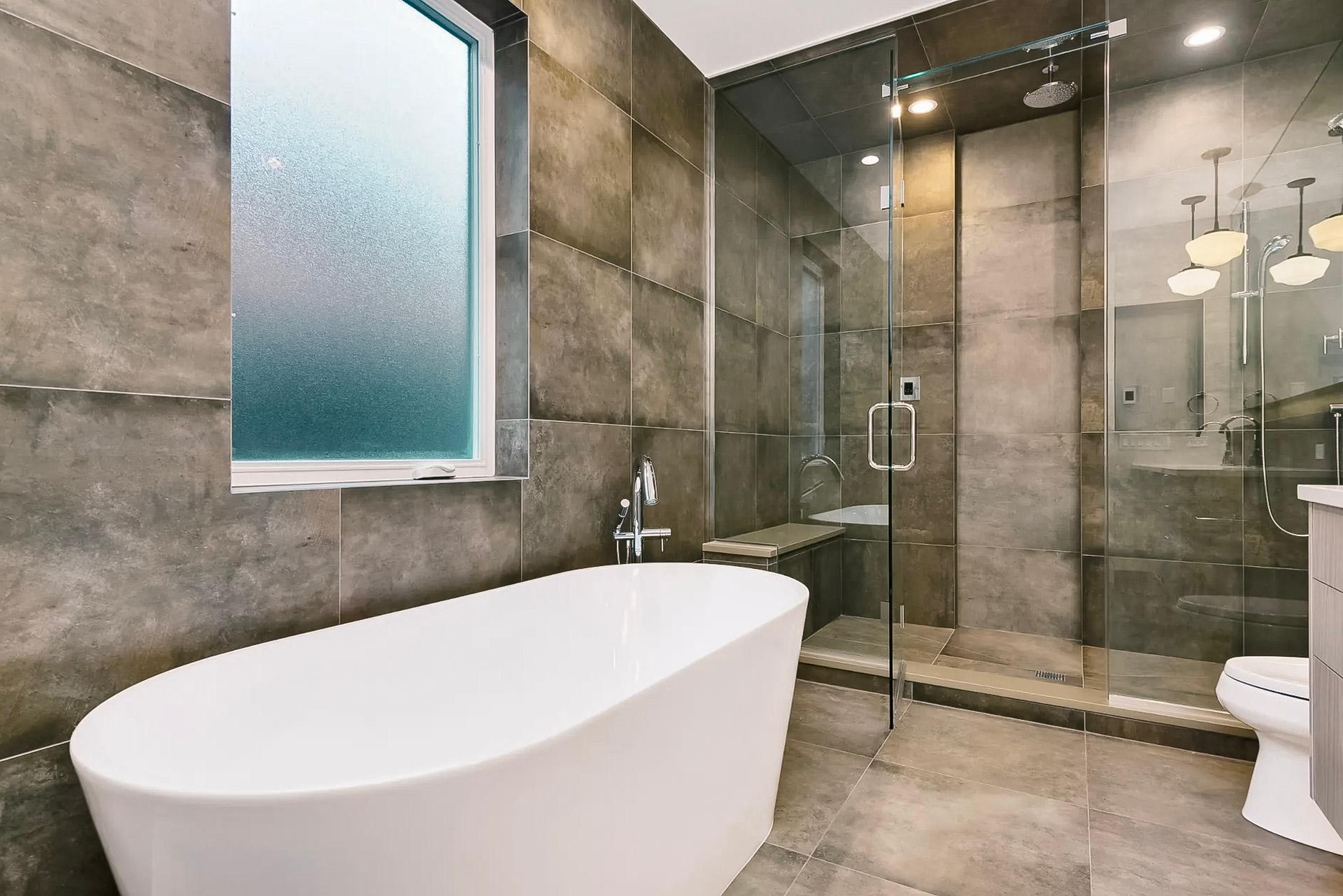 Salle de bain moderne avec baignoire autoportante blanche, douche en verre spacieuse et carrelage effet pierre aux murs et au sol. Fenêtre givrée pour plus d’intimité et robinetterie chromée.