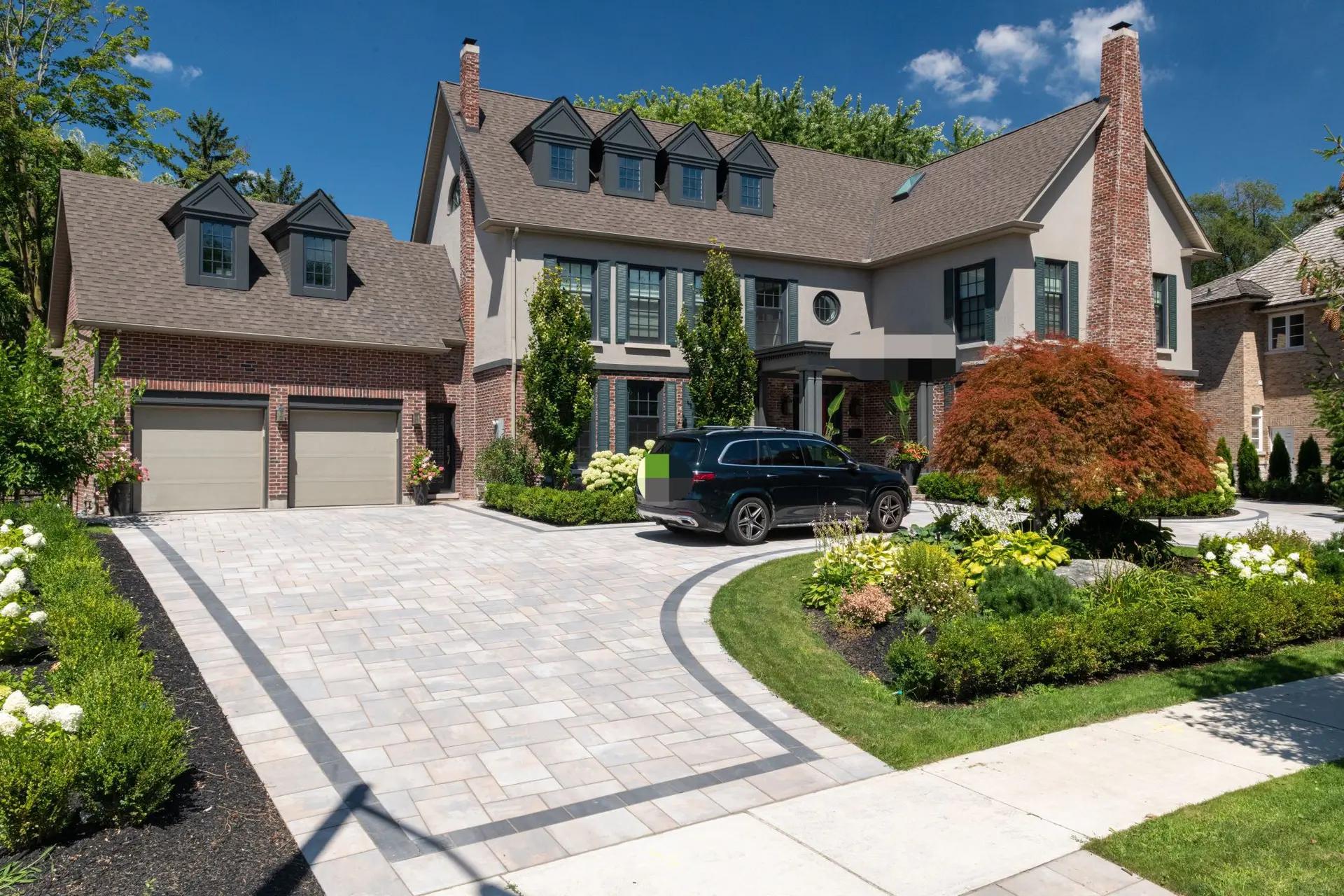 Grande maison de style traditionnel avec une entrée pavée spacieuse, un jardin paysager bien entretenu et une voiture stationnée devant le garage sous un ciel ensoleillé.