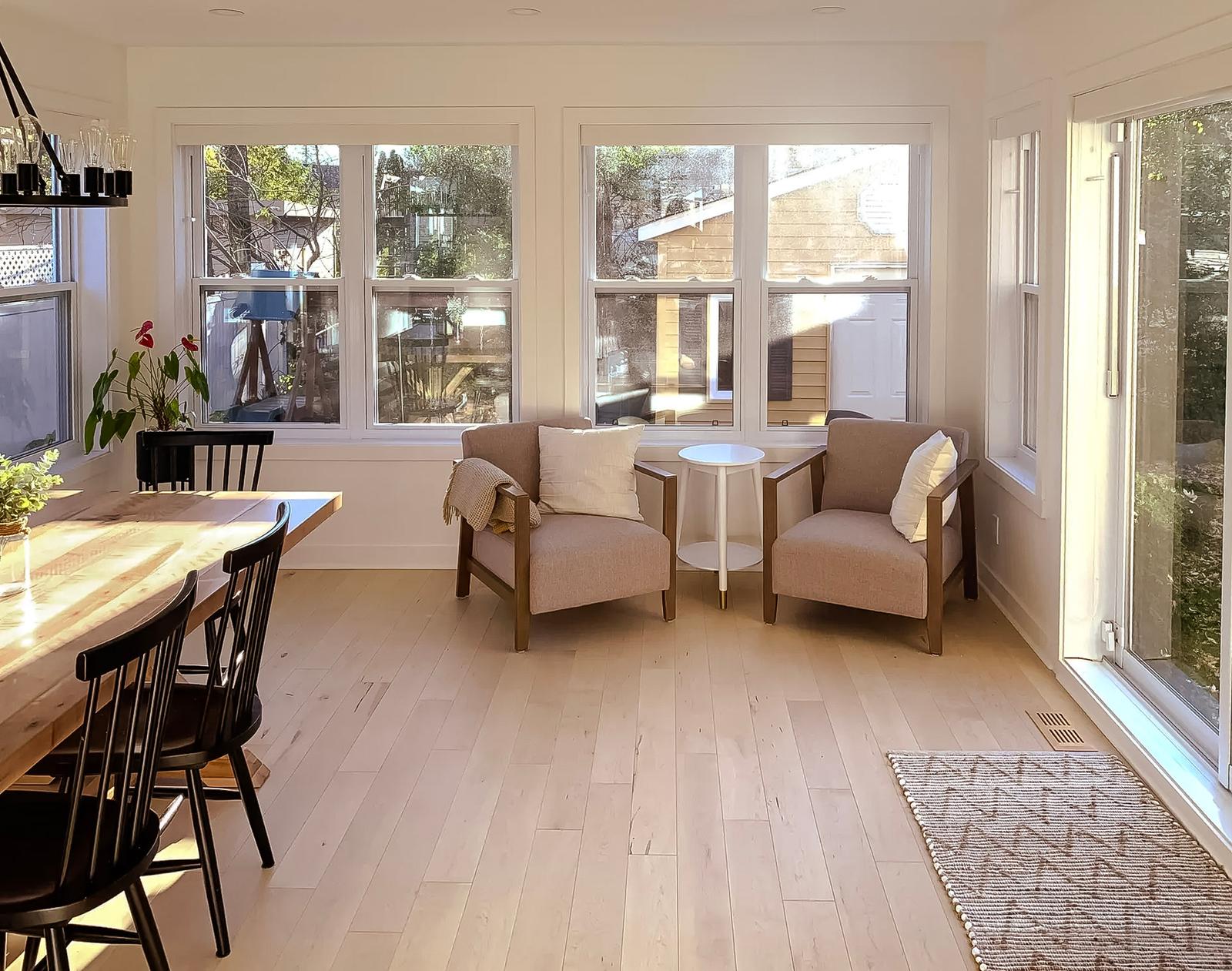 Salle à manger et coin salon lumineux avec grandes fenêtres, plancher en bois clair, table en bois massif et chaises noires. Deux fauteuils beiges avec coussins et table d'appoint blanche.