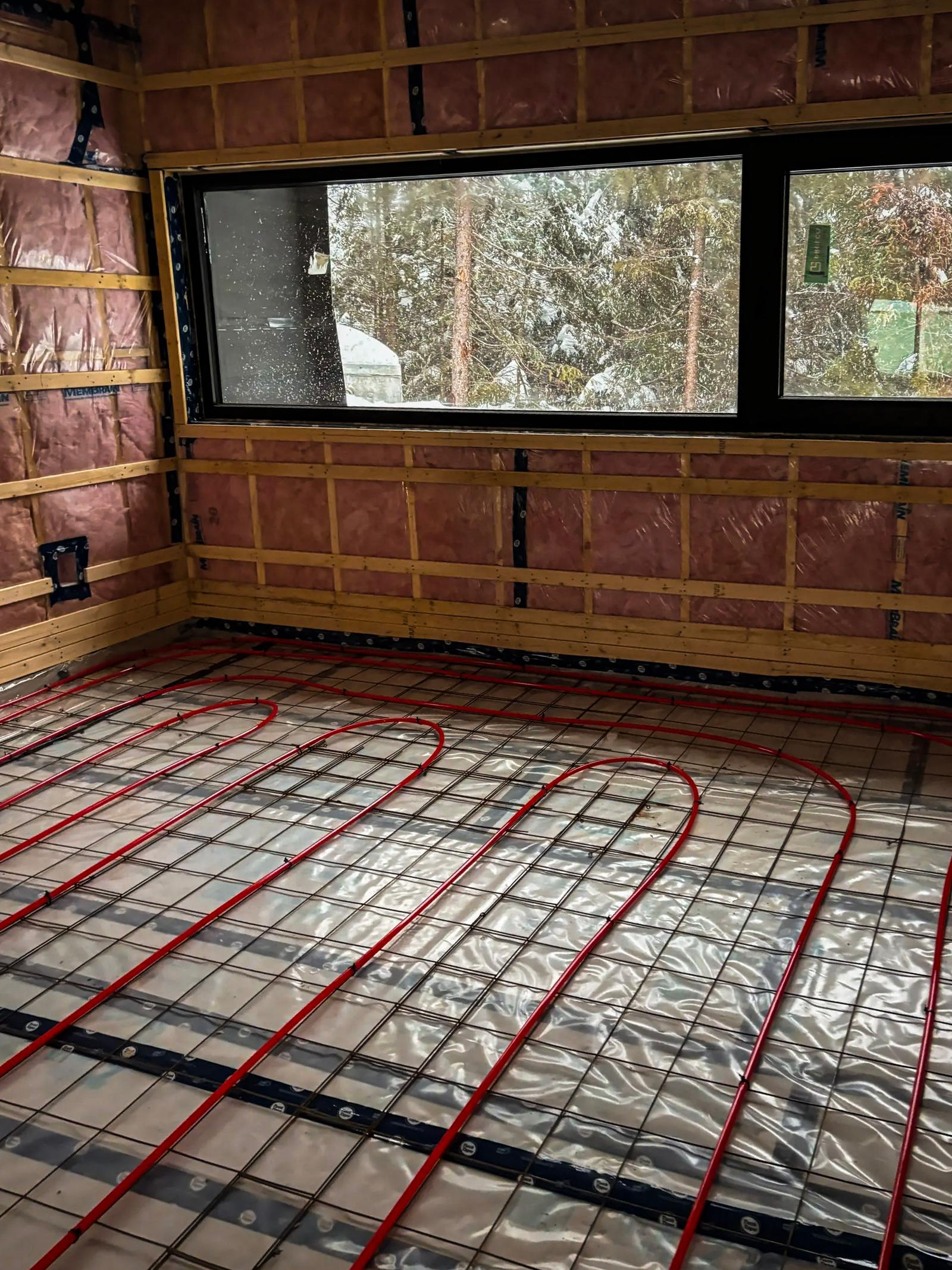 Système de chauffage au sol en cours d'installation dans une pièce en construction avec isolation et fenêtre panoramique.