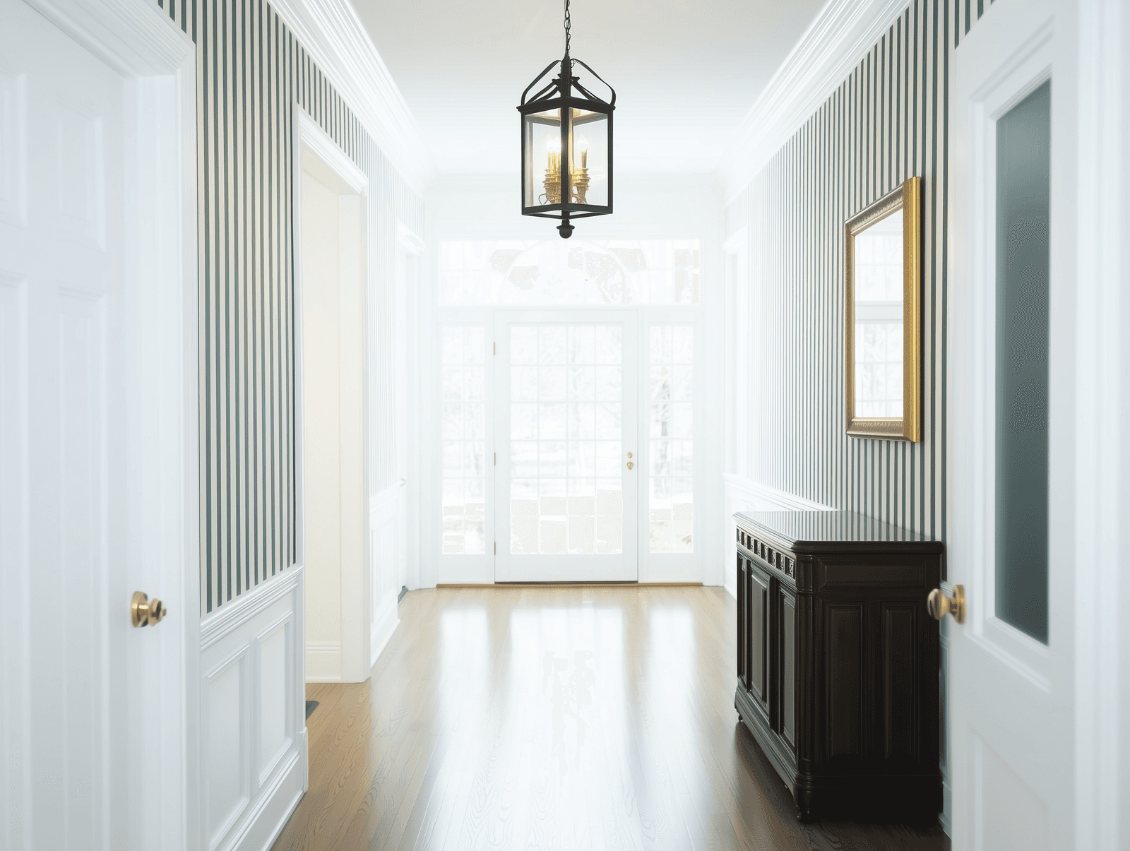 Entrée lumineuse et élégante avec murs à rayures, lustre suspendu, buffet en bois foncé et portes vitrées.