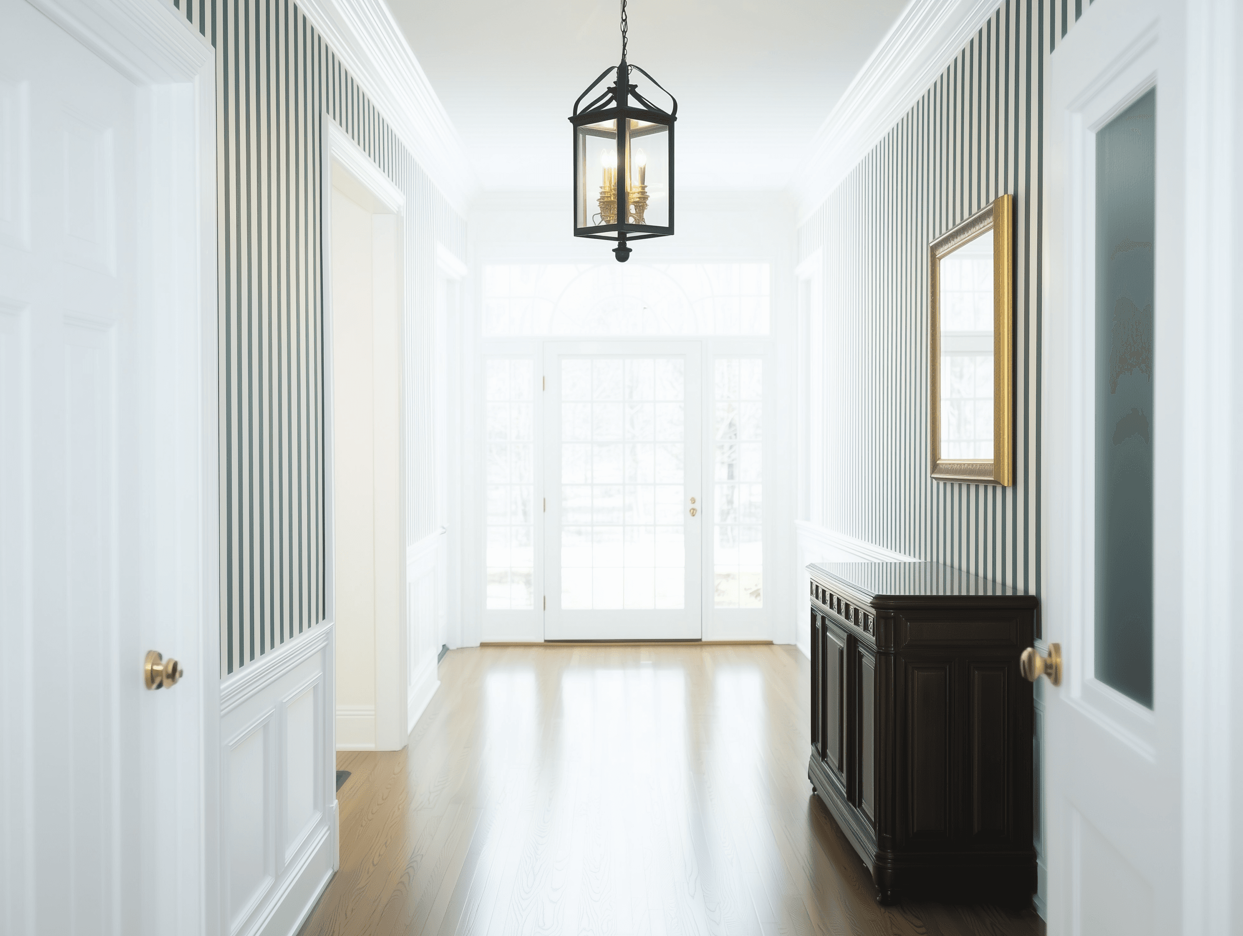 Entrée lumineuse et élégante avec murs à rayures, lustre suspendu, buffet en bois foncé et portes vitrées.