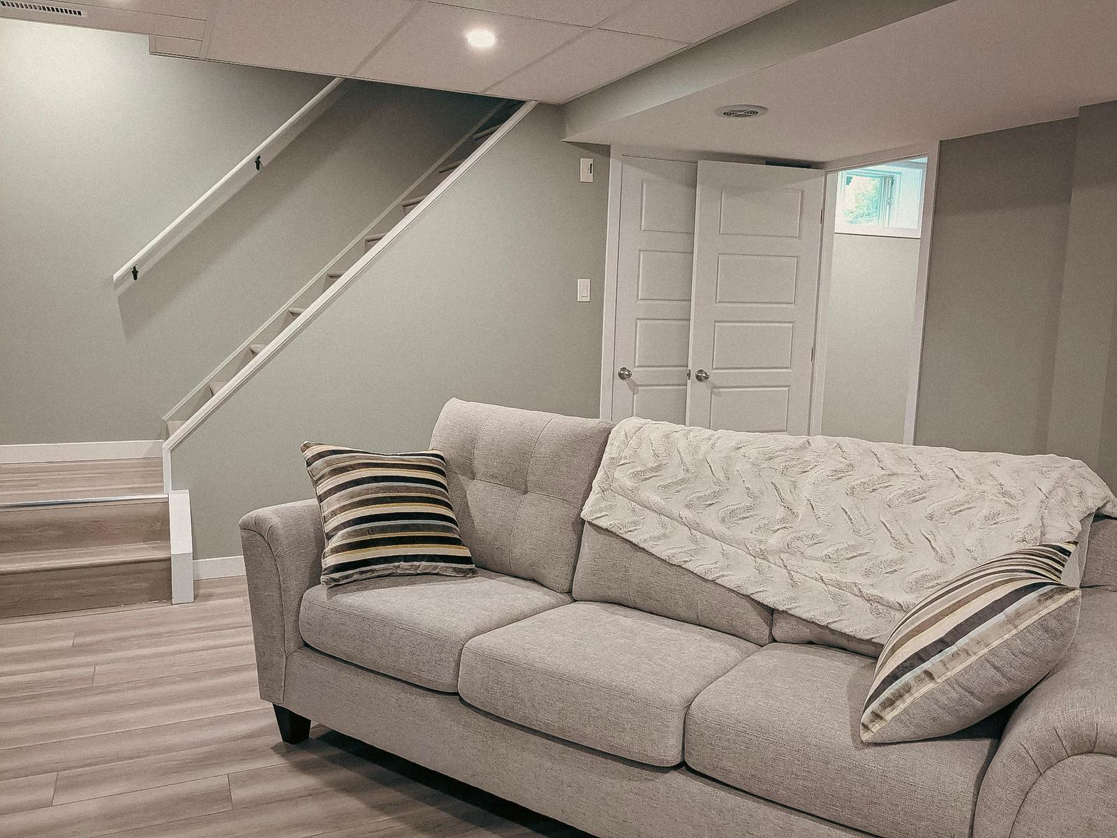 &nbsp;Sous-sol aménagé avec un canapé gris, des escaliers en bois et un mur neutre avec une porte blanche.