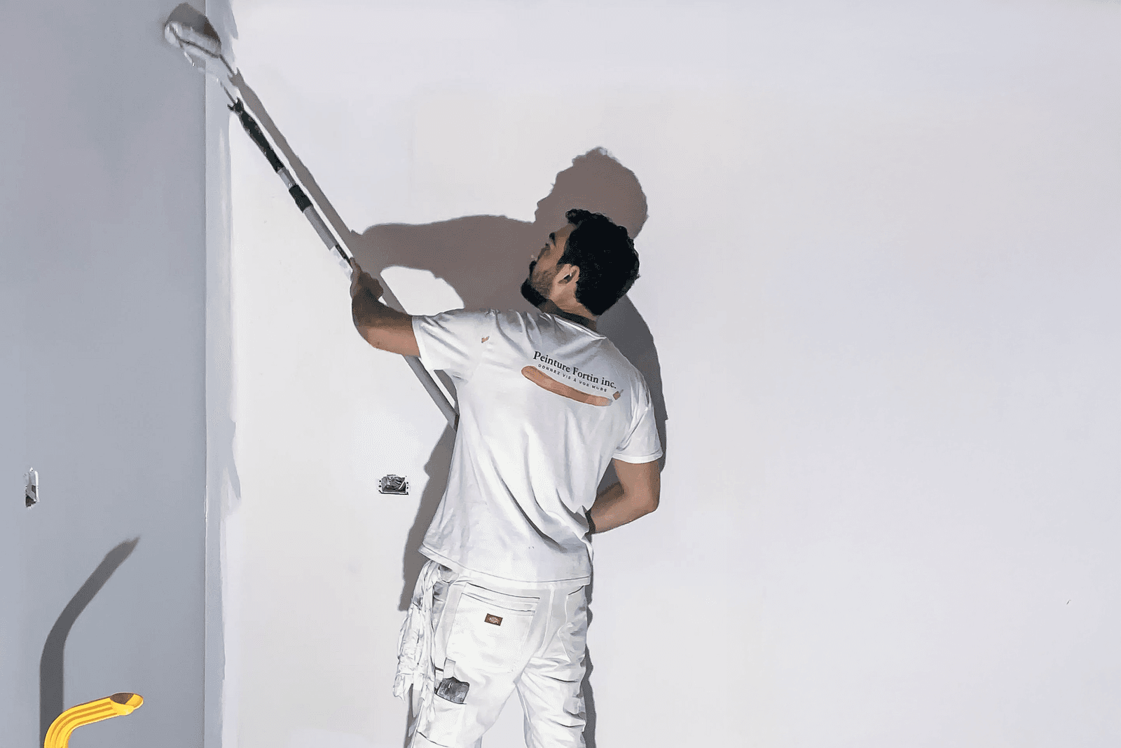 Peintre en bâtiment appliquant une couche de peinture blanche sur un mur à l’aide d’un rouleau télescopique, portant une tenue de travail blanche.