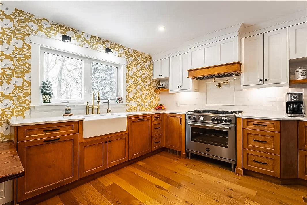 Cuisine rustique avec armoires en bois, papier peint floral jaune, évier farmhouse blanc, comptoirs en quartz blanc, et cuisinière en acier inoxydable.