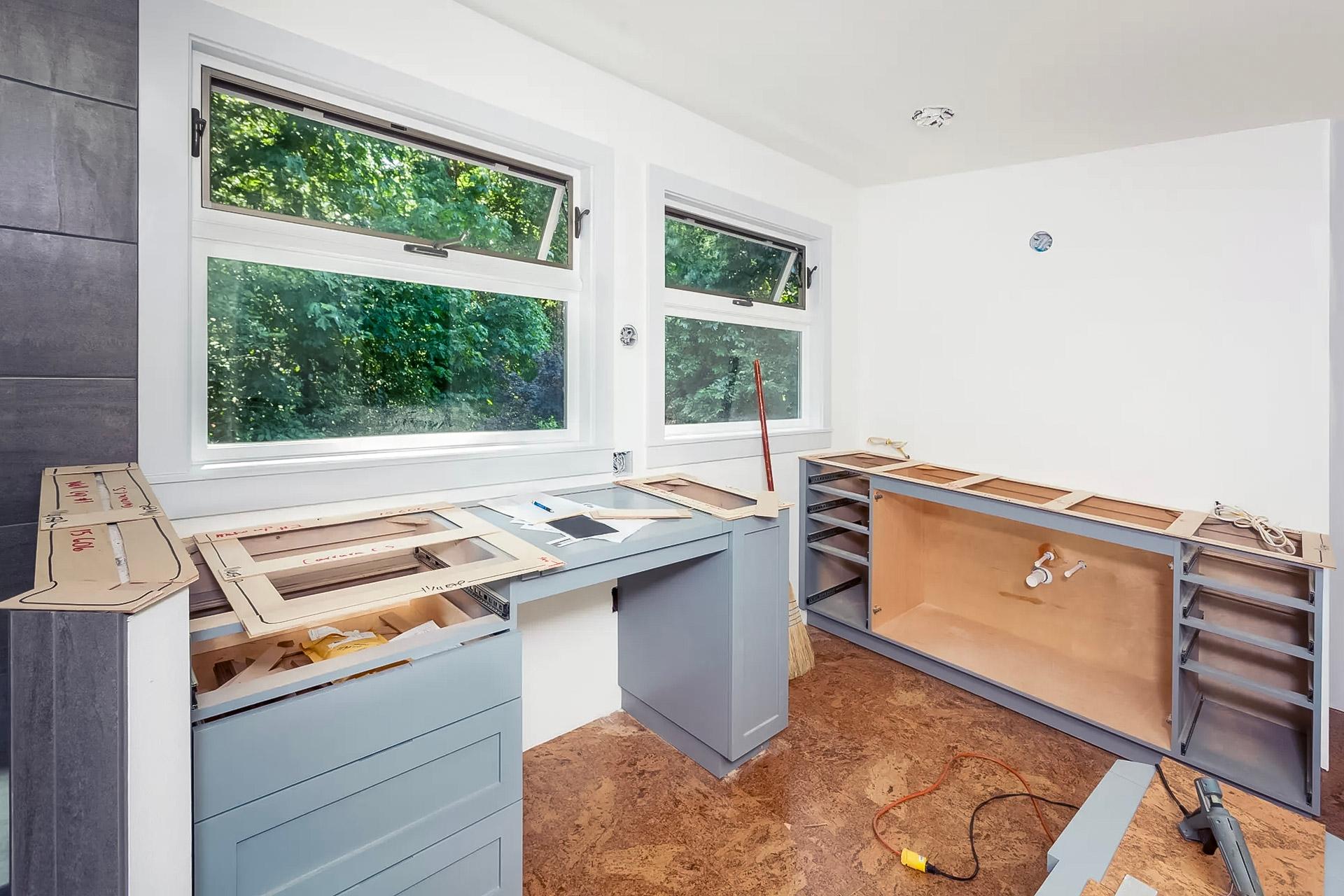 Cuisine en rénovation avec installation de nouveaux comptoirs et armoires grises sous des fenêtres lumineuses.