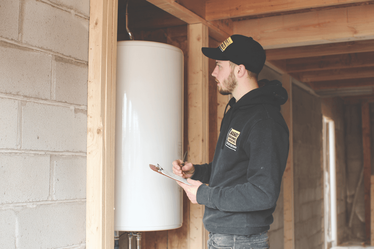 Un technicien en inspection portant une casquette et un chandail à capuchon noirs observe un chauffe-eau blanc dans une maison en construction, en prenant des notes sur une tablette.