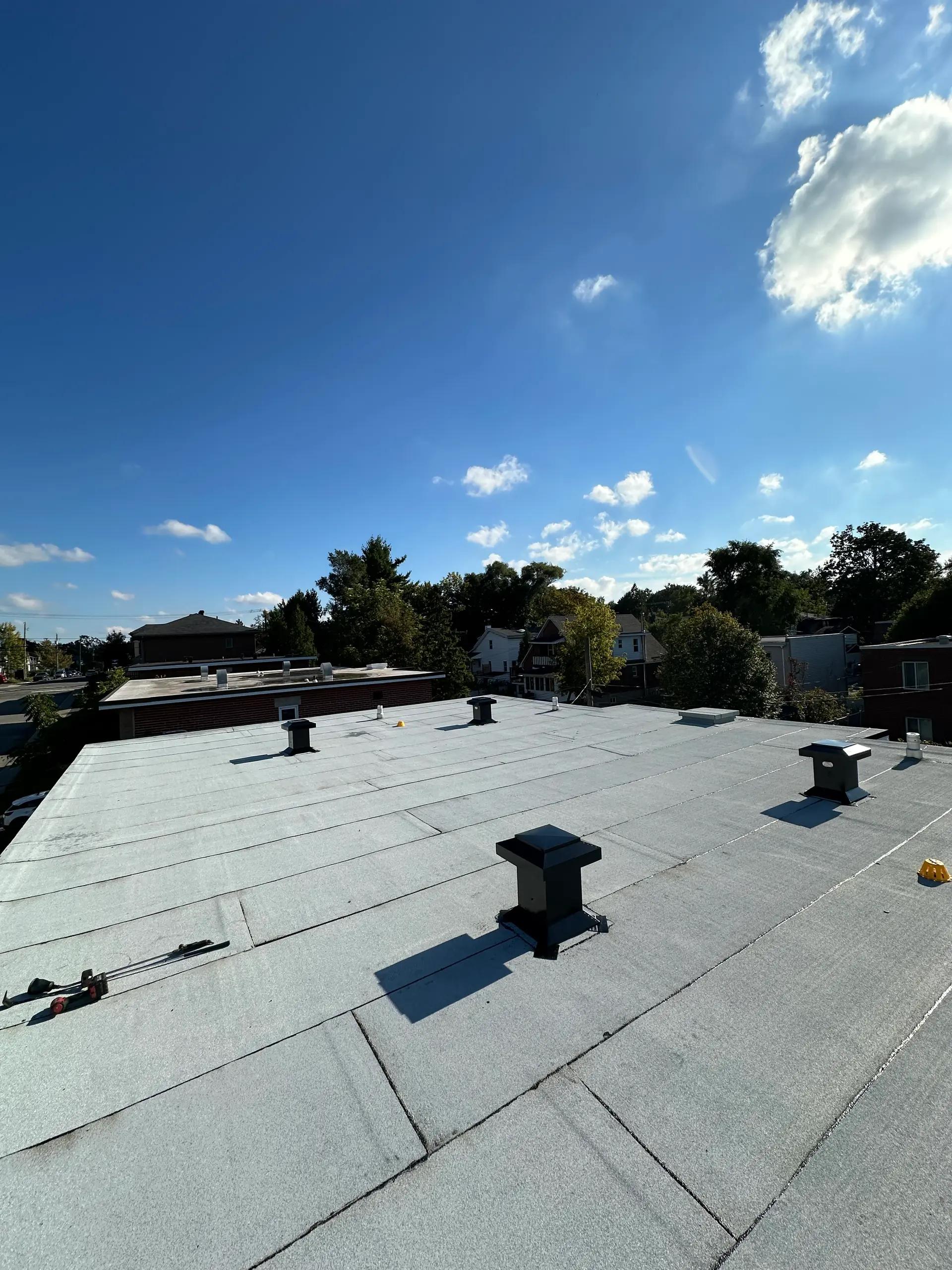 Toit plat recouvert d'une membrane grise avec plusieurs conduits de ventilation noirs, surplombant un quartier résidentiel verdoyant sous un ciel bleu dégagé.