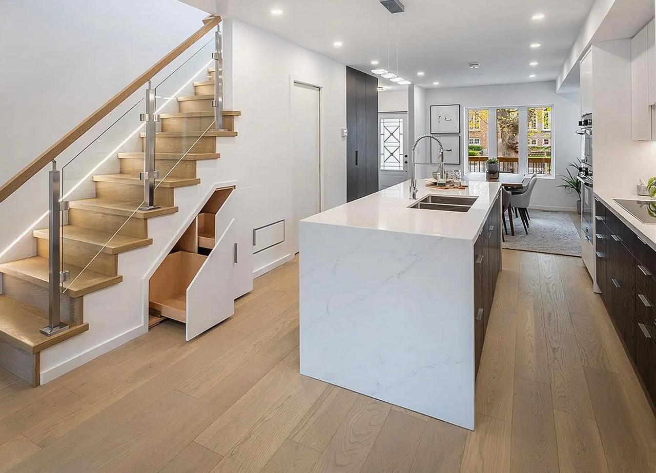 Intérieur de maison moderne avec cuisine ouverte, escalier en bois avec rangement intégré et îlot en marbre blanc. Vue sur la salle à manger lumineuse avec accès au jardin.