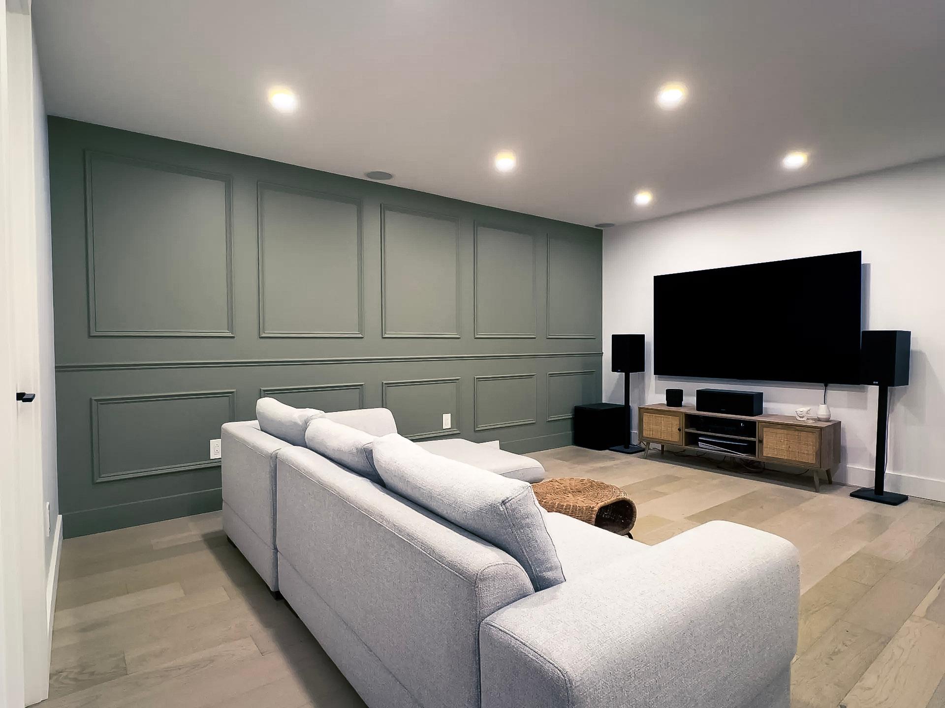 Salon moderne en sous-sol avec un mur accent vert, un grand canapé gris, un téléviseur écran plat et un système audio.