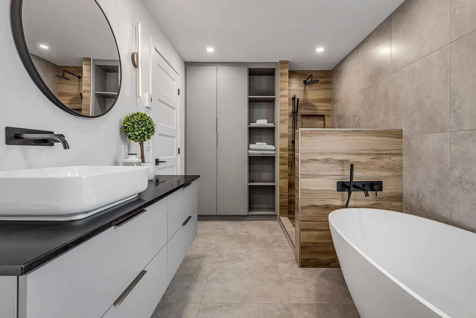 Salle de bain moderne avec baignoire autoportante, douche ouverte avec finition bois et murs en carrelage gris. Meuble-lavabo gris avec comptoir noir, vasque blanche et robinetterie noire mate.