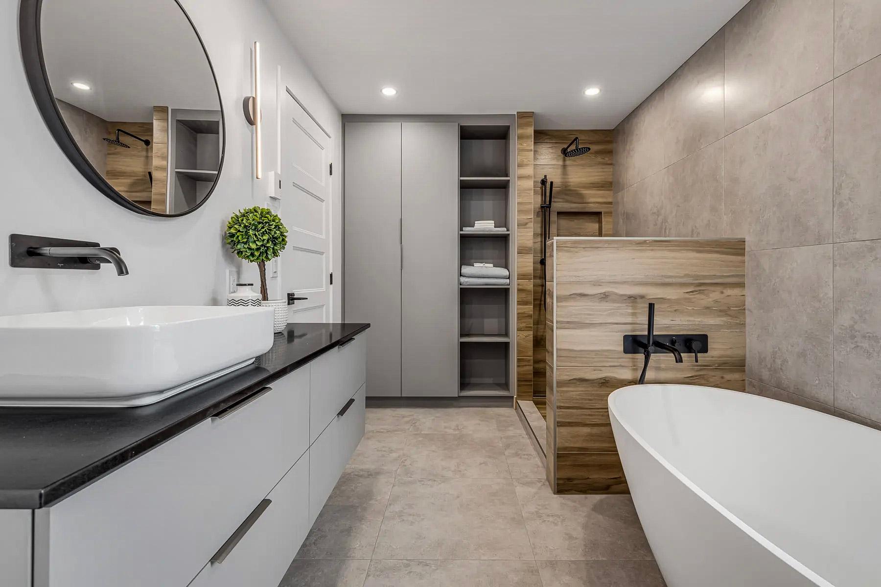Salle de bain moderne avec baignoire autoportante, douche ouverte avec finition bois et murs en carrelage gris. Meuble-lavabo gris avec comptoir noir, vasque blanche et robinetterie noire mate.