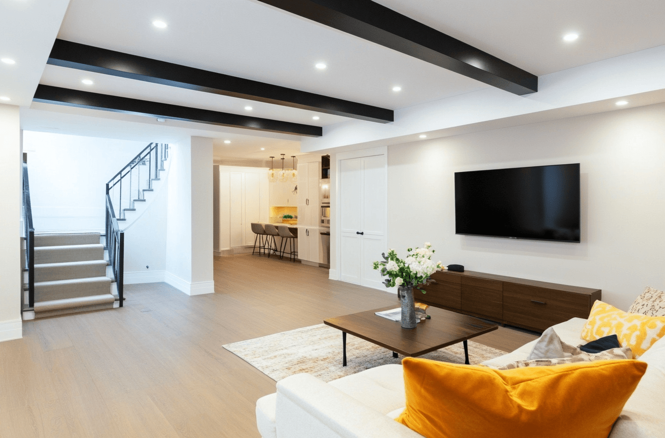Salon moderne et lumineux avec poutres noires au plafond, canapé blanc avec coussins jaunes, télé murale, escalier élégant et coin repas avec éclairage suspendu.