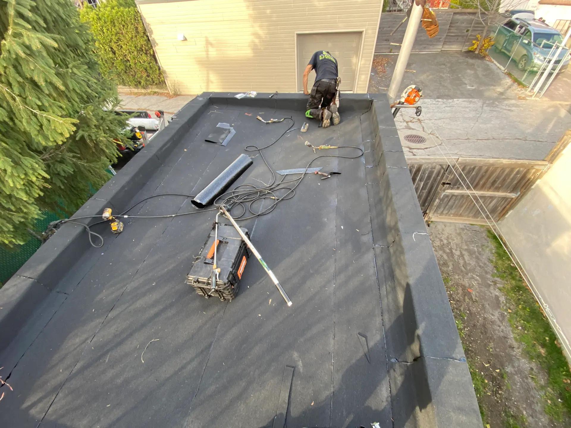 Travaux d'installation d'une nouvelle membrane noire sur un toit plat, avec un ouvrier en pleine pose, entouré d'outils et d'équipements dans une cour résidentielle clôturée