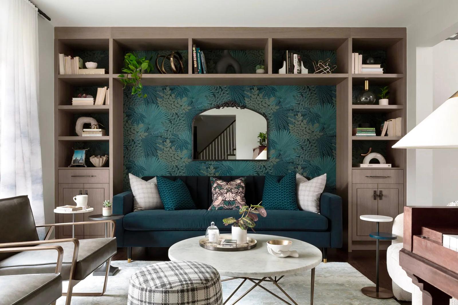 Salon moderne avec canapé en velours bleu, papier peint floral, miroir design, bibliothèque intégrée et table basse en marbre dans un espace lumineux et élégant.