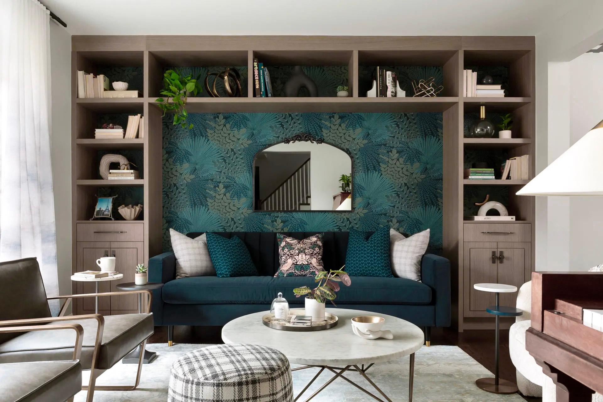 Salon moderne avec canapé en velours bleu, papier peint floral, miroir design, bibliothèque intégrée et table basse en marbre dans un espace lumineux et élégant.