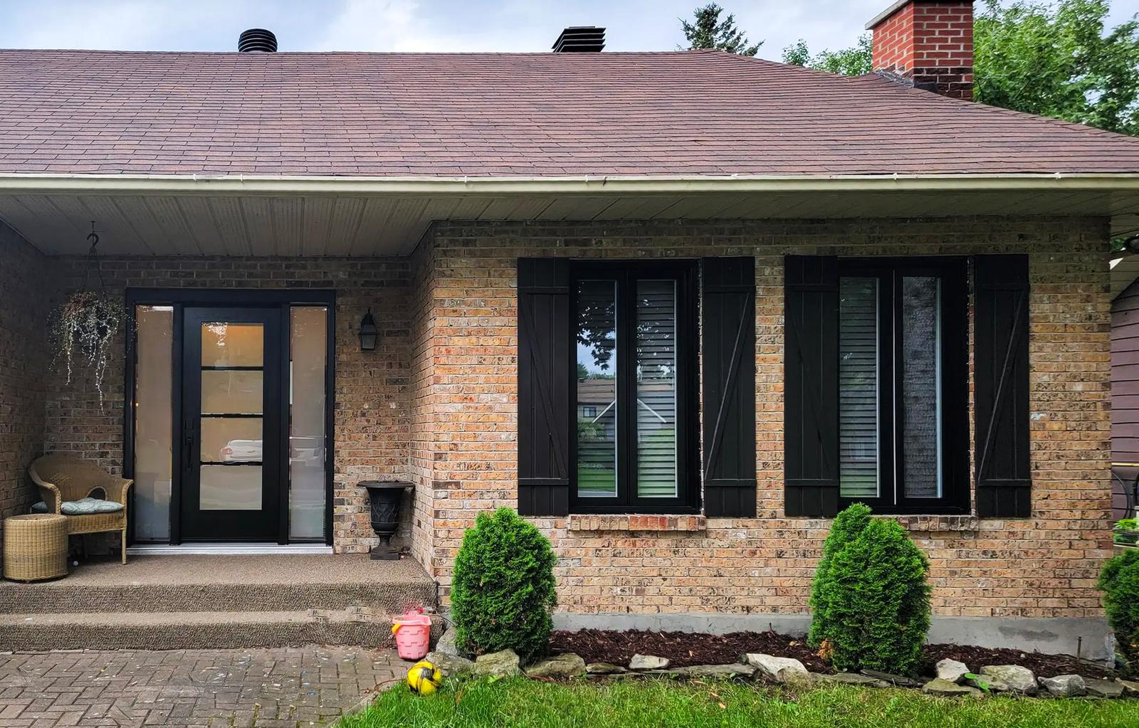 Maison en brique avec porte noire vitrée, volets assortis et petit aménagement paysager en façade.<br>