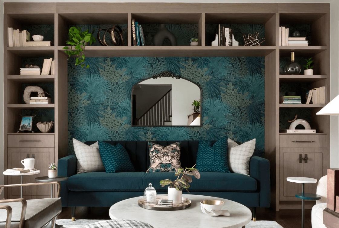 Salon moderne avec canapé en velours bleu, papier peint floral, miroir design, bibliothèque intégrée et table basse en marbre dans un espace élégant.