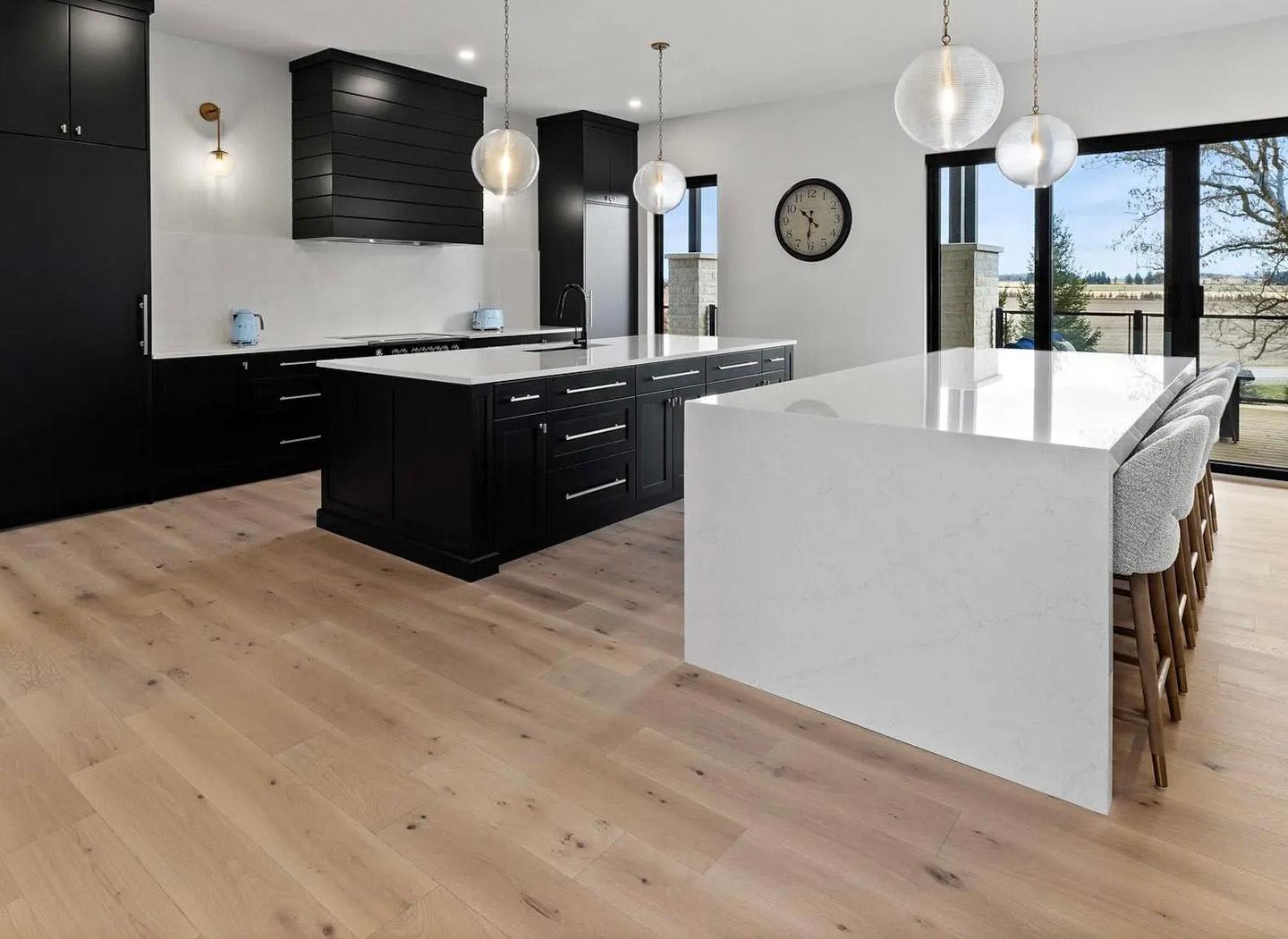 Cuisine moderne avec armoires noires, îlot en marbre blanc et plancher en bois clair. Éclairage suspendu en verre, grandes portes vitrées donnant sur l'extérieur et tabourets rembourrés.