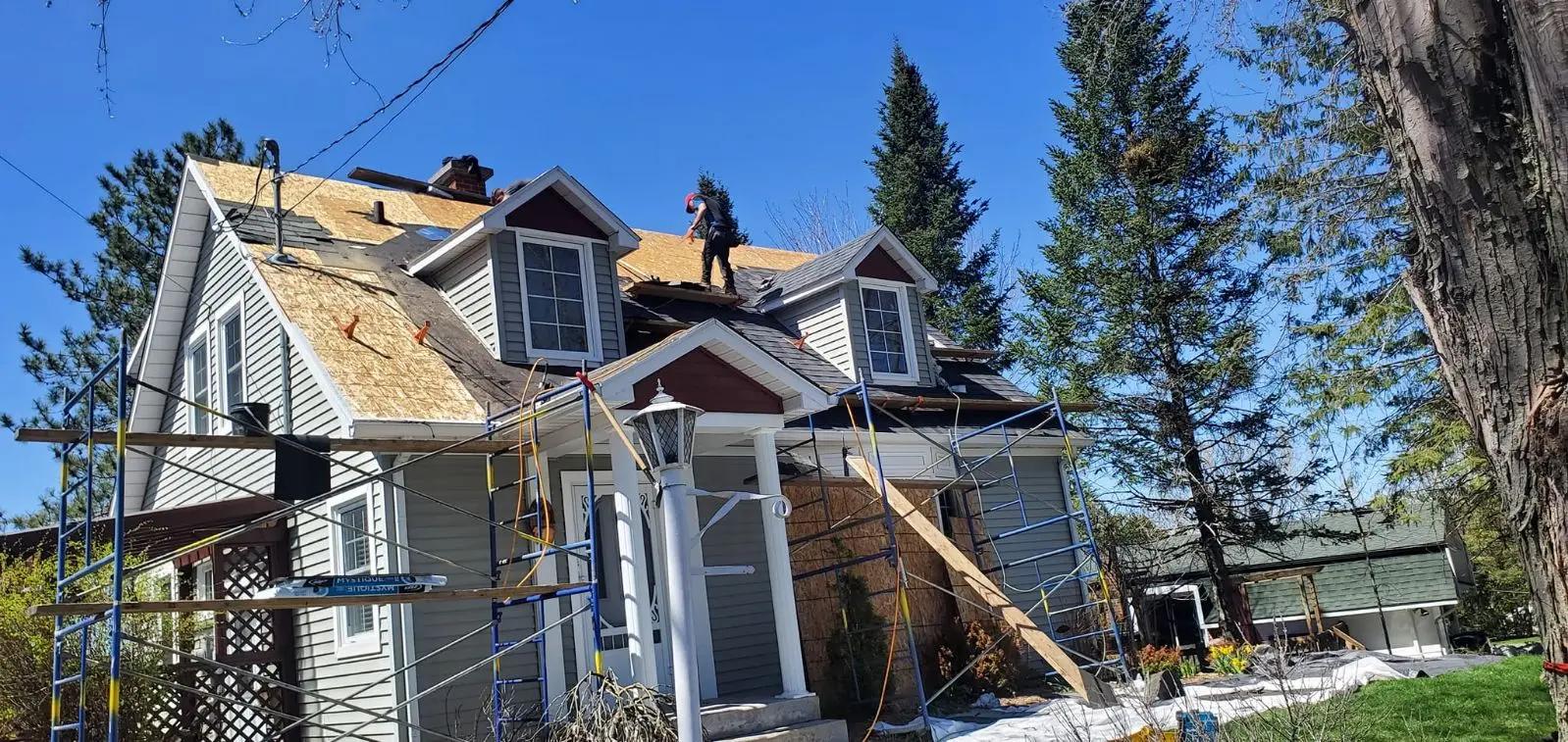 Maison en rénovation avec des échafaudages autour et des ouvriers travaillant sur la toiture, en train de poser des panneaux de contreplaqué sous un ciel dégagé.