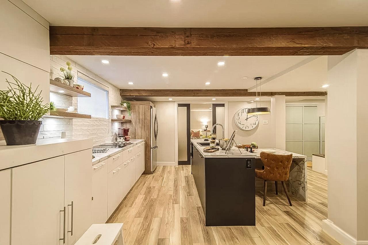 Cuisine moderne avec îlot central, poutres en bois au plafond, armoires blanches, éclairage encastré et sol effet bois