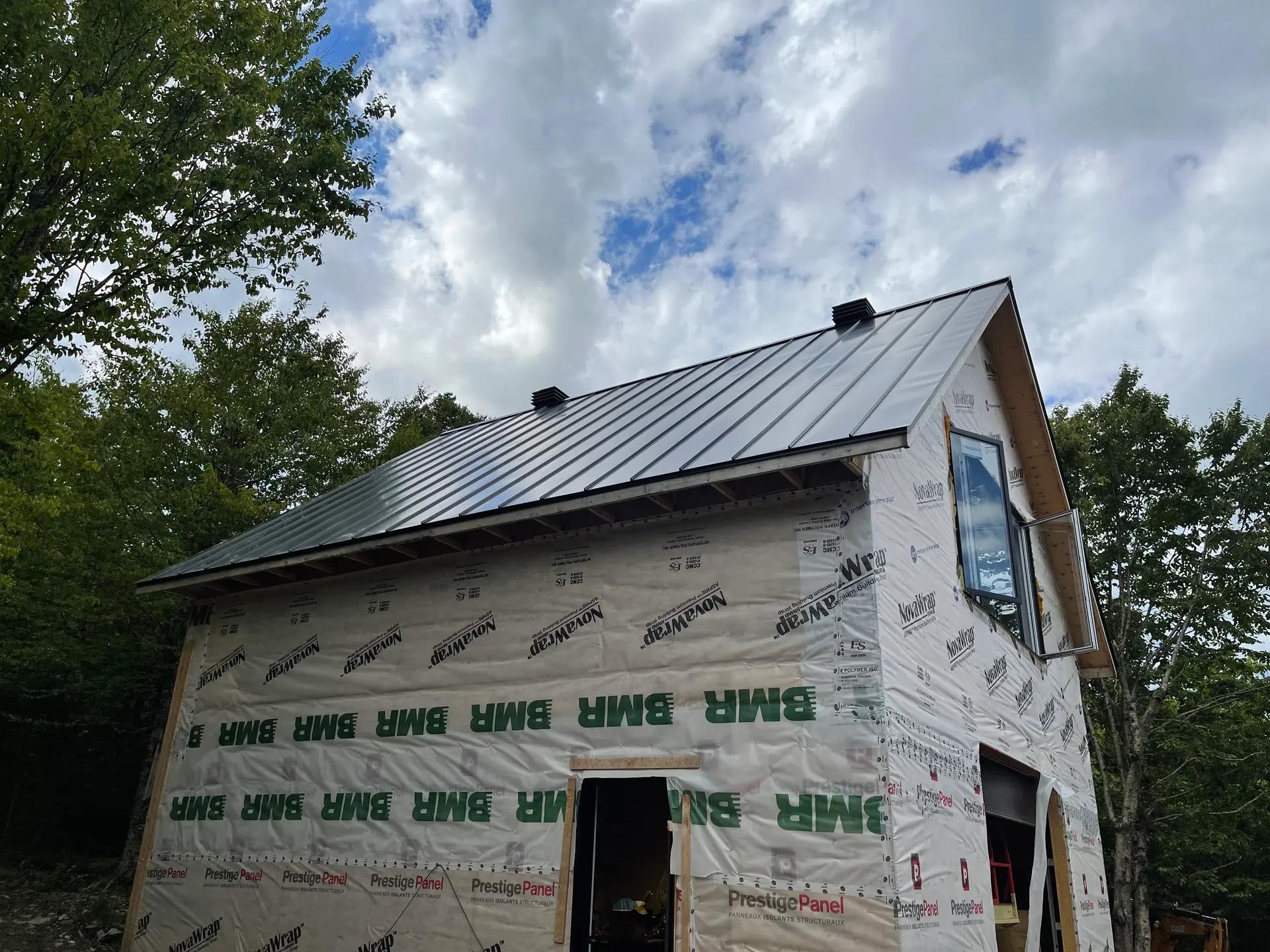 Bâtiment résidentiel en cours de construction, avec un toit en métal gris installé. Les murs sont encore couverts de matériaux de protection et d'isolants temporaires portant les logos 'BMH' et 'PrestigePanel'. Située dans un cadre naturel entouré d'arbres, la structure est conçue avec un toit à pignon et une fenêtre à l'étage. Le ciel nuageux ajoute une atmosphère de chantier en plein air.