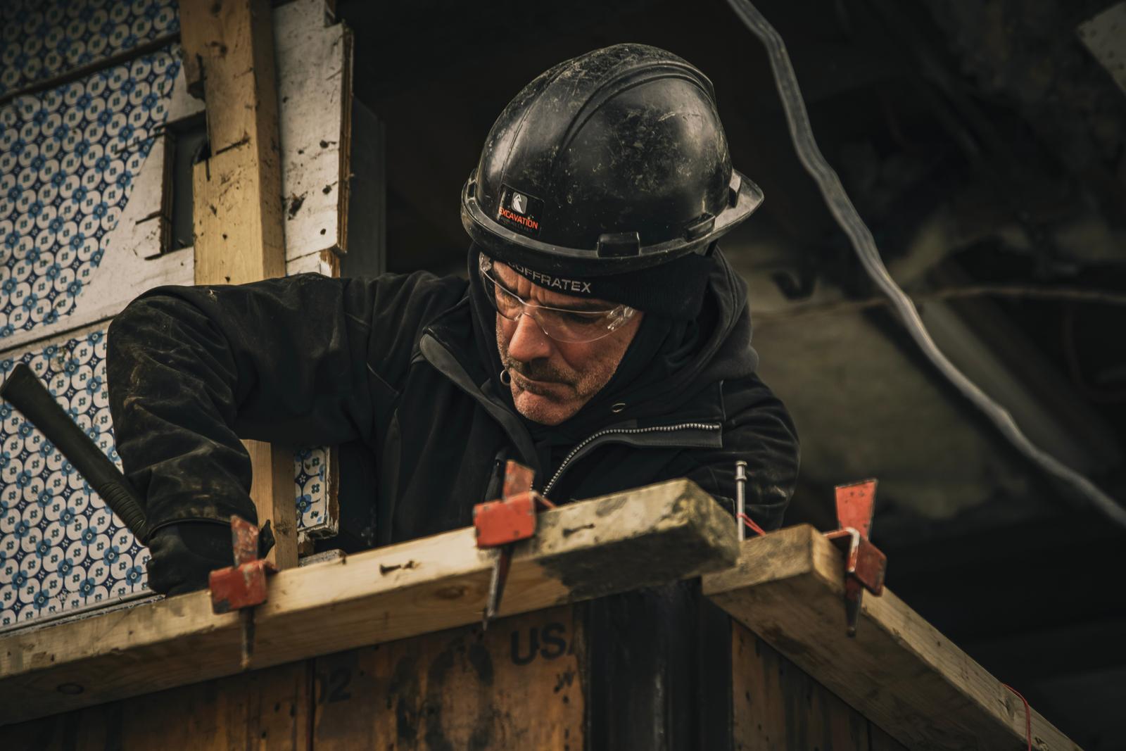 Travailleur portant un casque noir et des lunettes de protection, effectuant des travaux de menuiserie et de construction sur un chantier.
