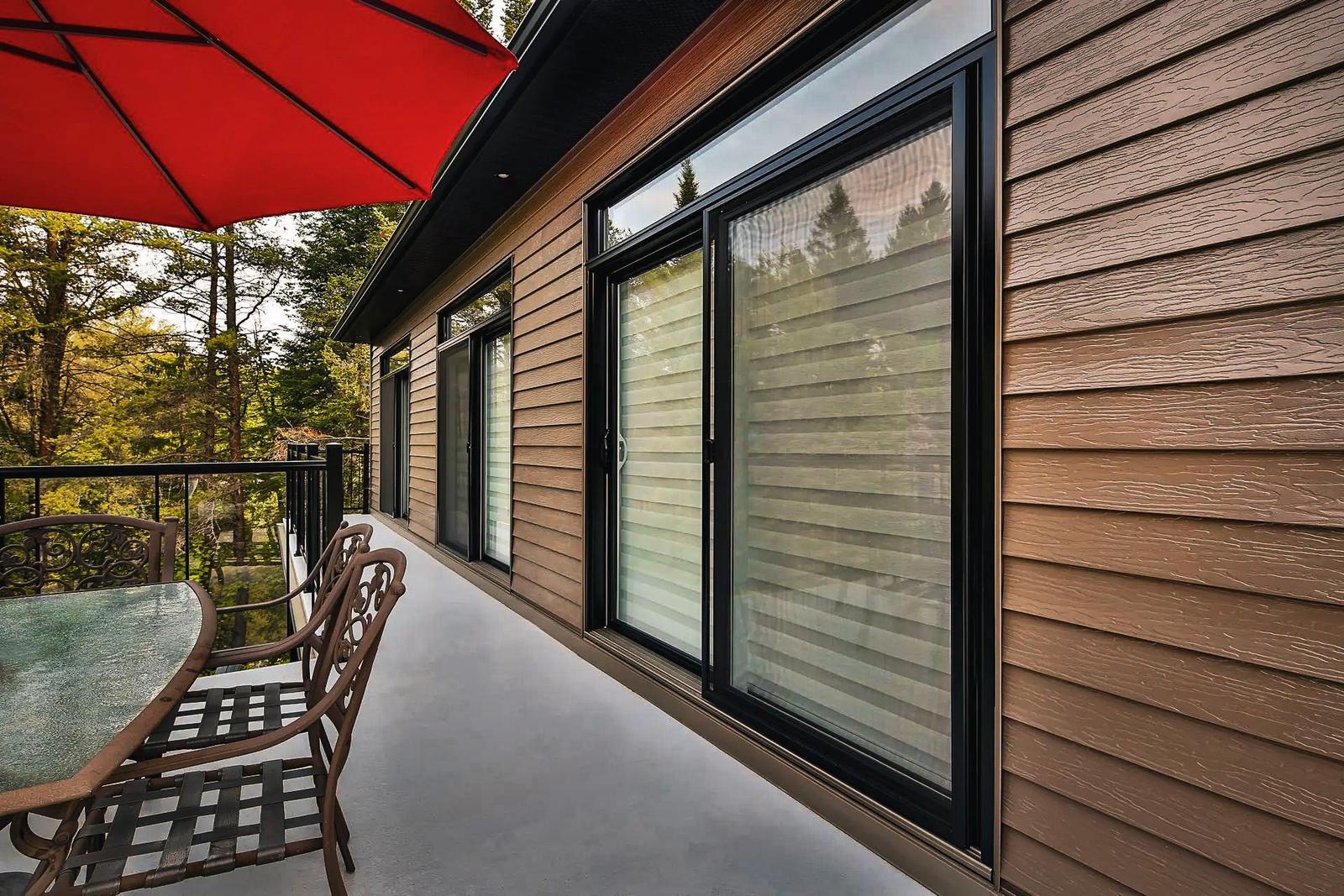 Terrasse extérieure moderne avec revêtement en bois, grandes fenêtres coulissantes et mobilier de terrasse en métal sous un parasol rouge.