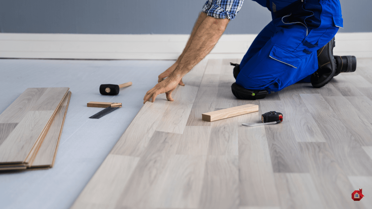 Pose d’un plancher flottant en bois clair par un rénovateur à genoux, avec outils de pose visibles (marteau, règle, cales, ruban à mesurer)