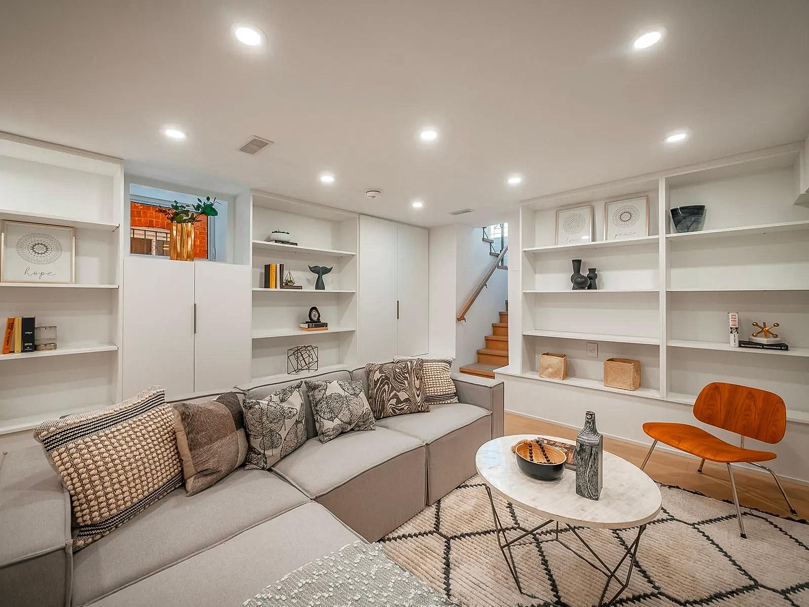Sous-sol aménagé avec salon moderne comprenant un canapé modulaire, une bibliothèque intégrée, une table basse en marbre, un fauteuil en bois et une décoration minimaliste.